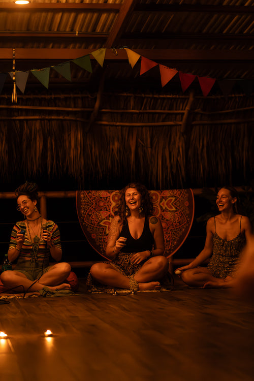Three women sitting on the floor, laughing and smiling, with a banner of flags and candles, representing the joyful and communal atmosphere of the "Sol y Luna" retreat.