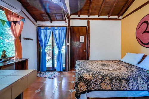 A cozy guest room at the Vida Asana retreat center, featuring a bed with a patterned comforter, a spiritual-themed wall painting, and a door leading to a balcony.