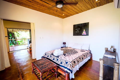 A bright and airy bedroom at a tropical retreat with a bed featuring a patterned blanket, a bamboo ceiling, and a doorway leading to a balcony.